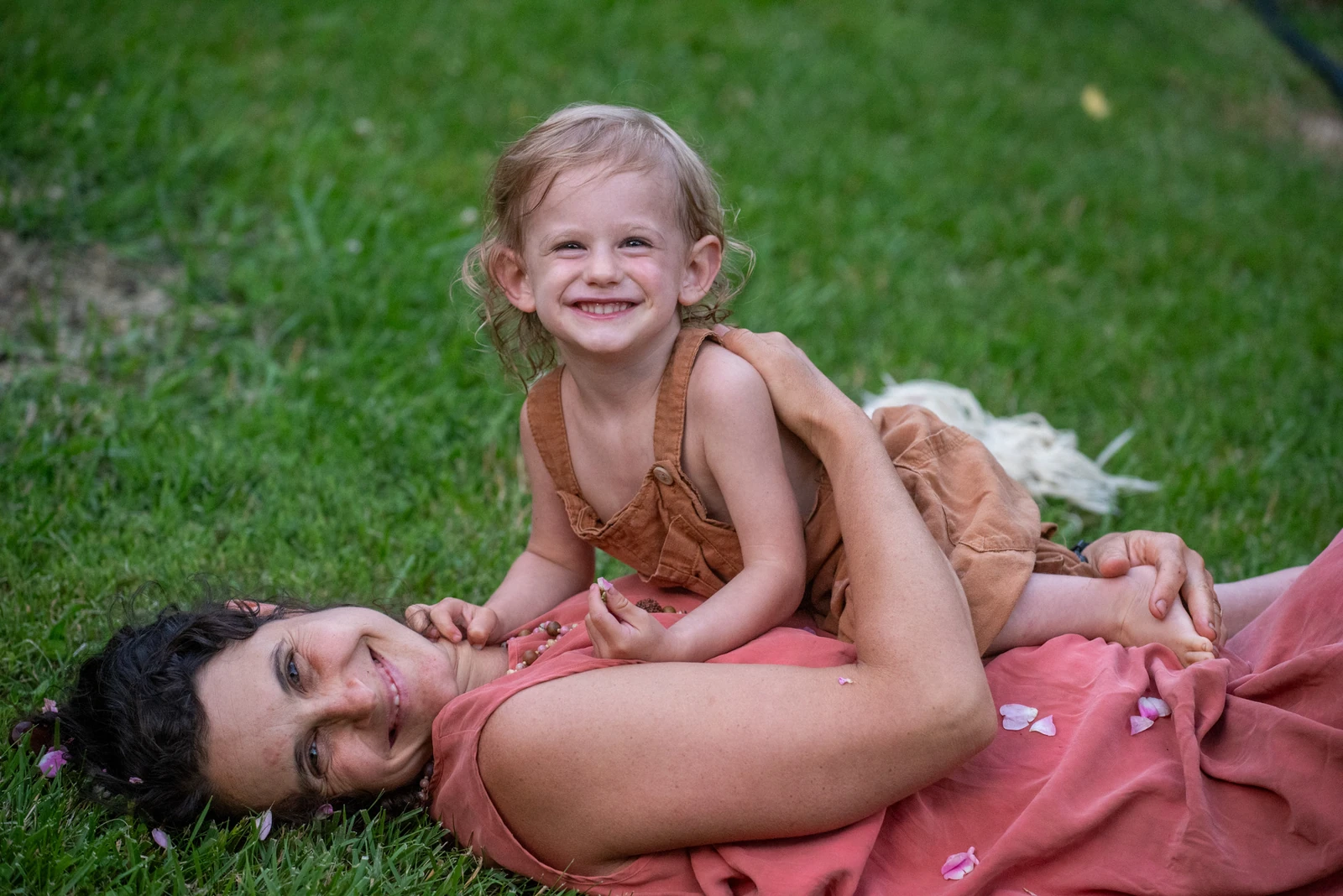 A mother laughing with her toddler while lying together in the grass, surrounded by flower petals